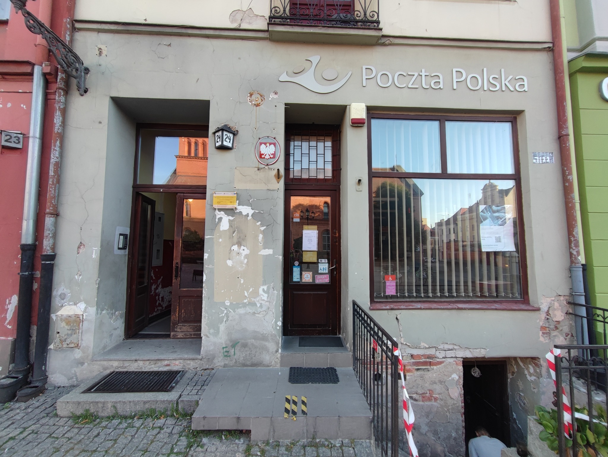 Wejście do budynku Poczty Polskiej z widocznymi śladami łuszczącej się farby na elewacji i uszkodzeniami tynku, podkreślającymi potrzebę renowacji.