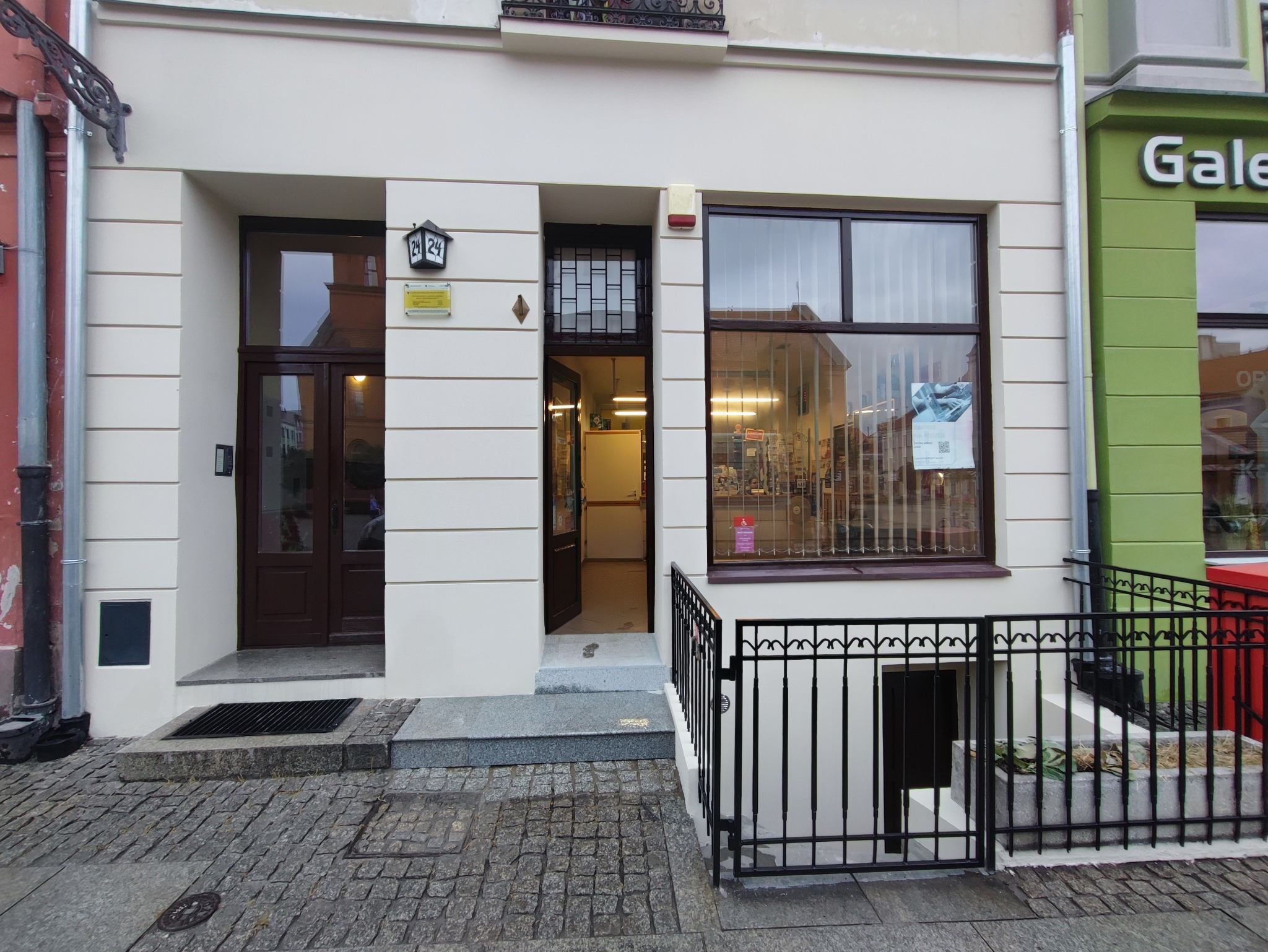 Odnowiona elewacja budynku z jasną fasadą, ciemnymi ramami okiennymi i drzwiami wejściowymi, widok na wnętrze przez otwarte drzwi, numer budynku 24 nad wejściem.