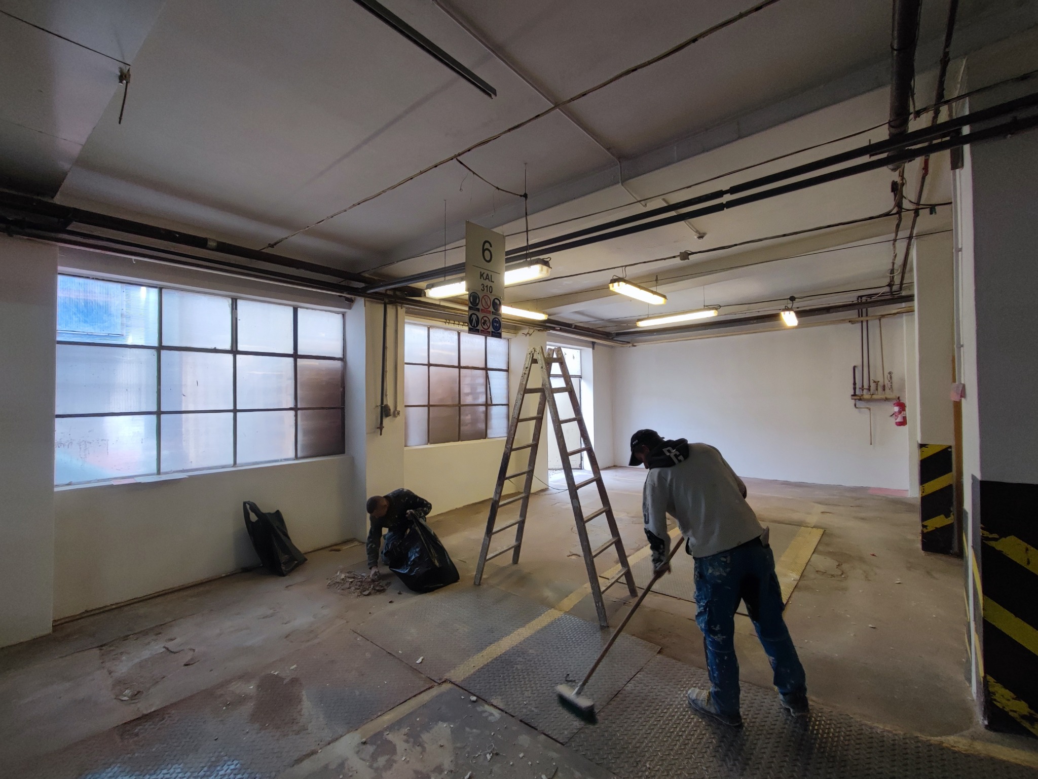 Dwóch pracowników sprząta podłogę po remoncie w przestronnym pomieszczeniu z dużymi oknami i drabina na środku, jeden zbiera gruz do czarnego worka, drugi zamiata podłogę szczotką.