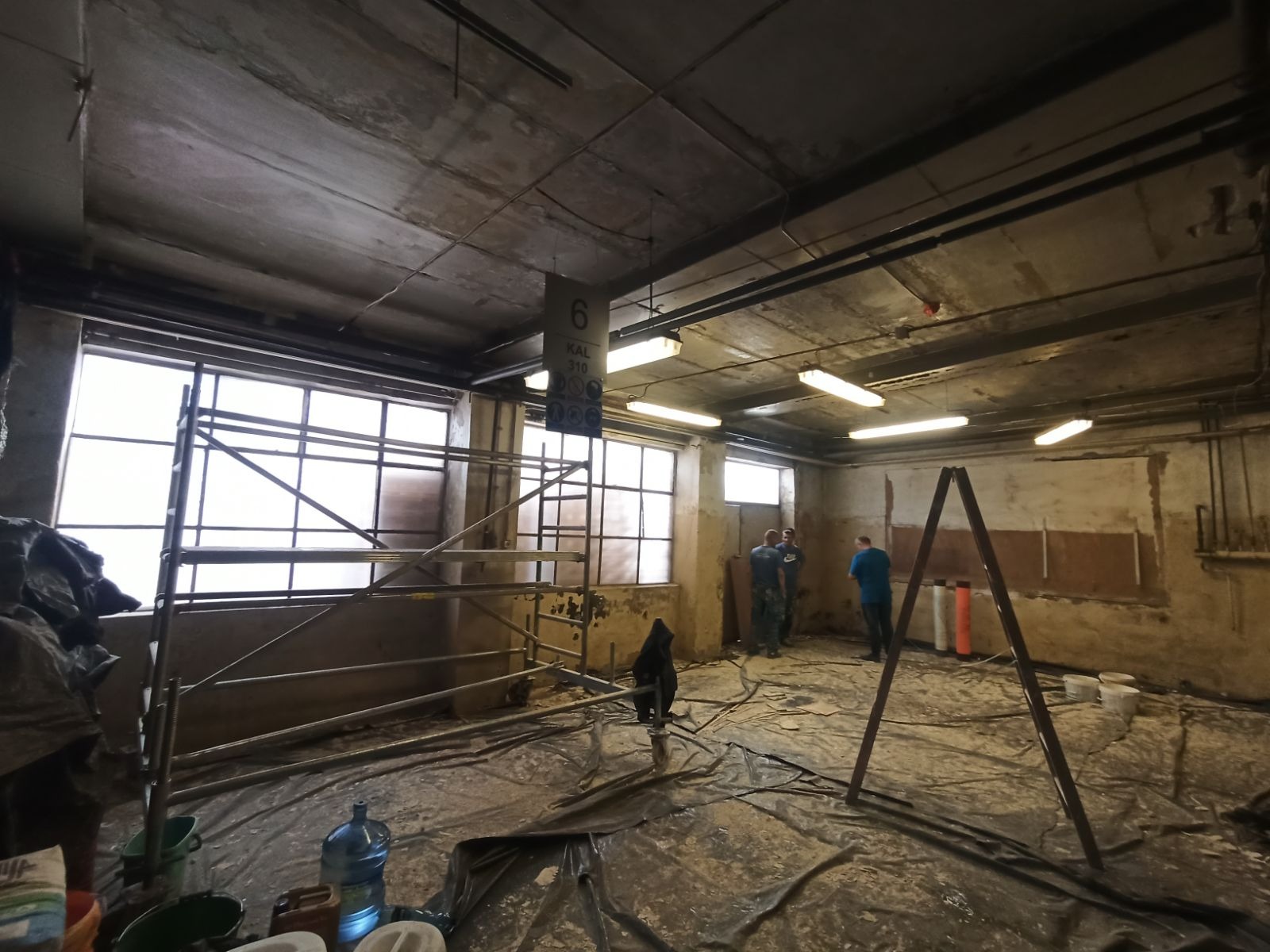 Wnętrze w trakcie remontu, zabezpieczona folią podłoga, rusztowania przy oknach, widoczni trzej mężczyźni w tle, surowy betonowy sufit z lampami.