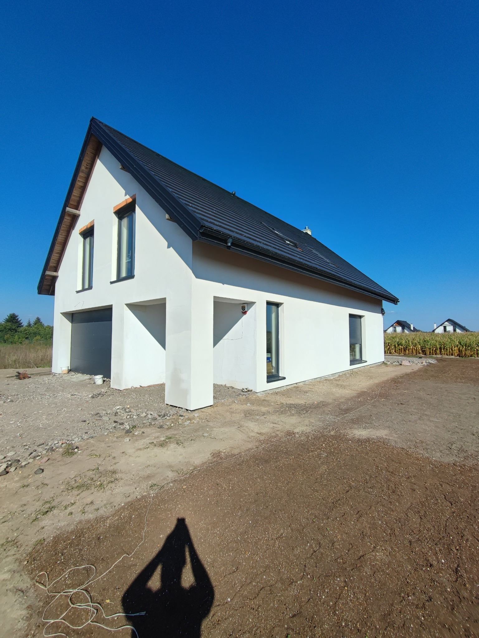 Nowoczesny, biały dom jednorodzinny z czarnym dachem, sfotografowany szerokokątnie w słoneczny dzień, z widocznym cieniem fotografa na pierwszym planie.