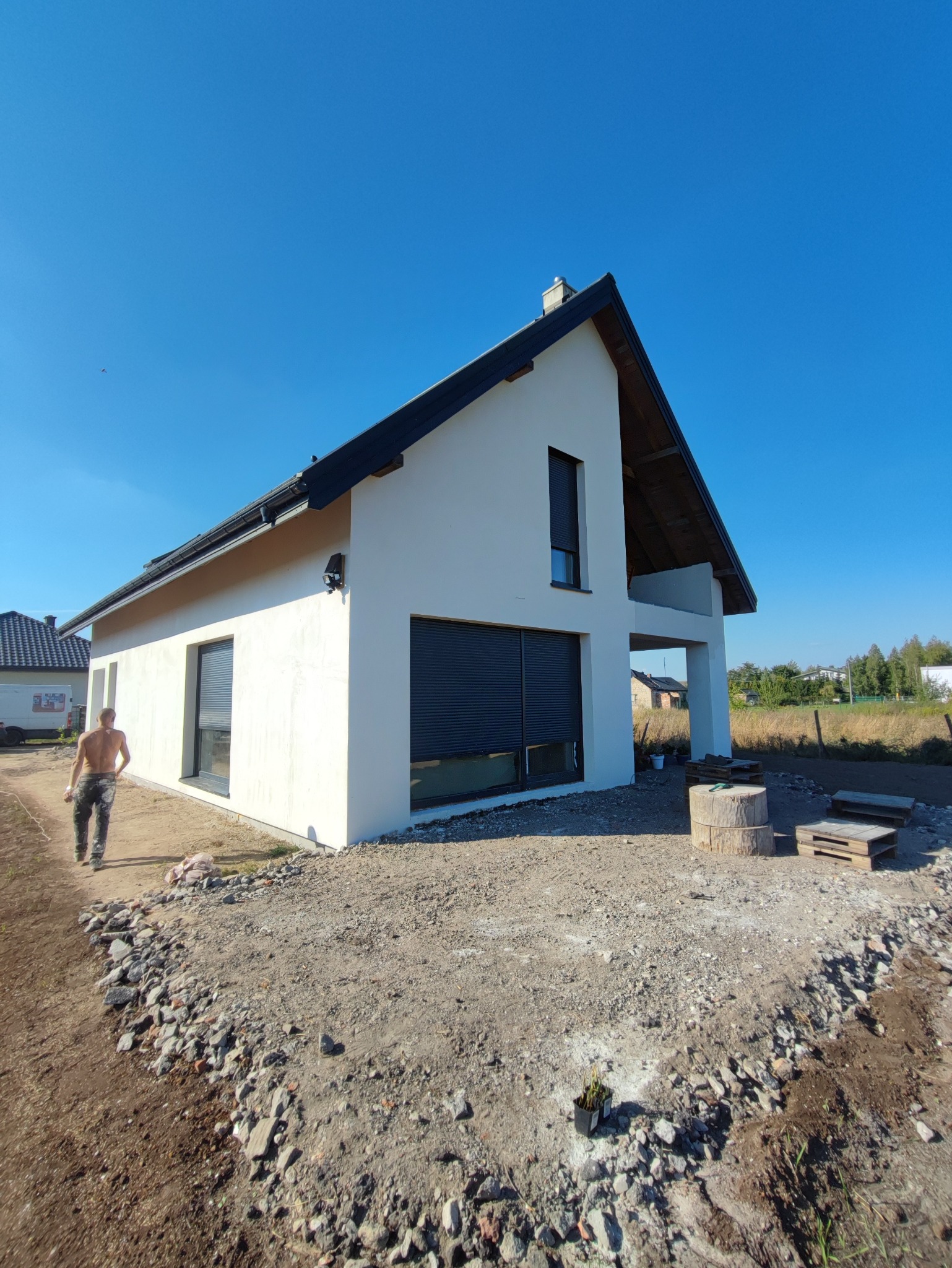 Nowoczesny dom jednorodzinny z białą elewacją i czarnymi ramami okiennymi, widoczne niedokończone prace ziemne wokół budynku i mężczyzna w oddali.