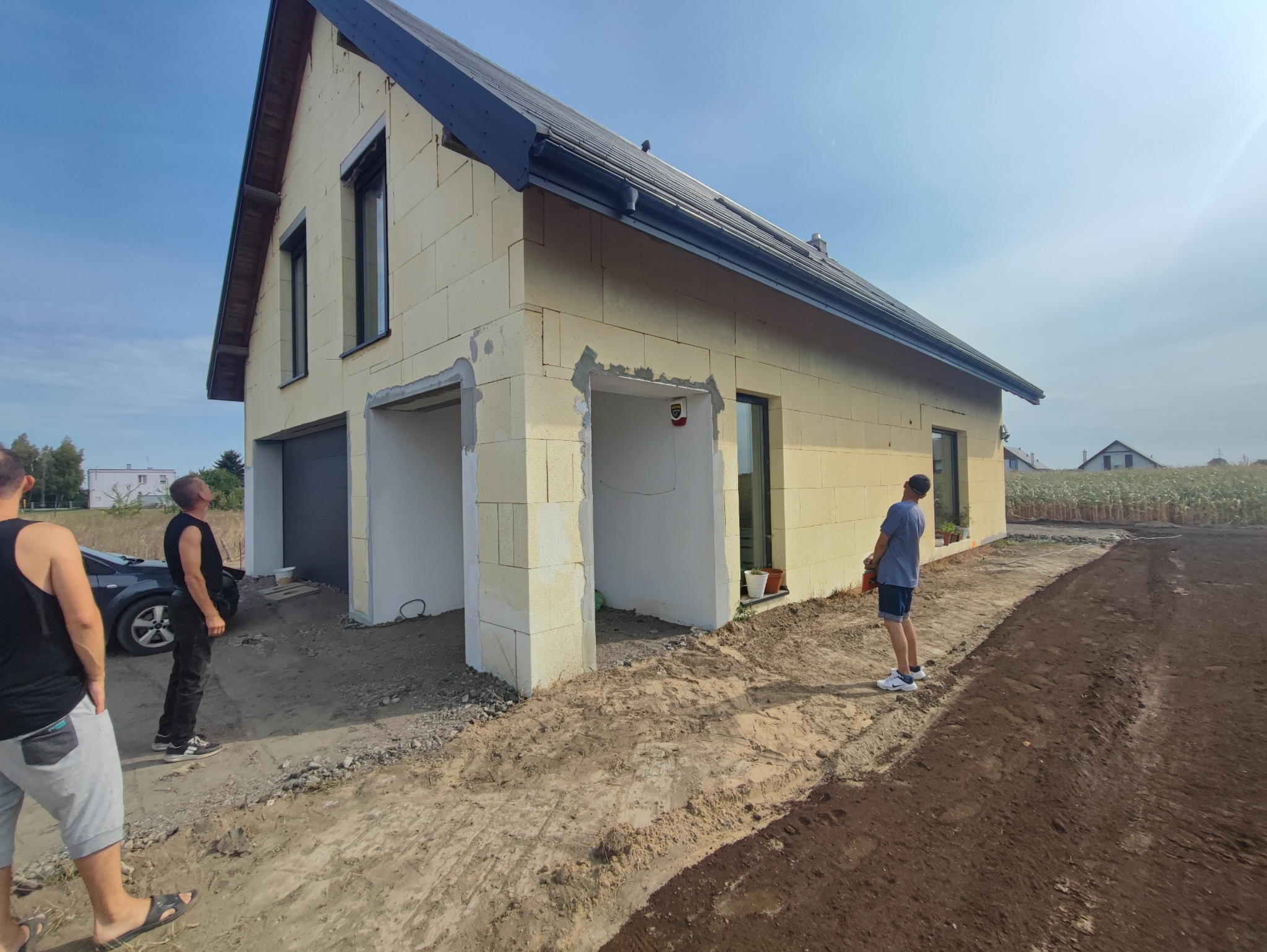 Surowy stan domu jednorodzinnego z pustaków, z widocznymi otworami na okna i drzwi, niezabezpieczonym terenem wokół i grupą osób obserwujących postęp prac.