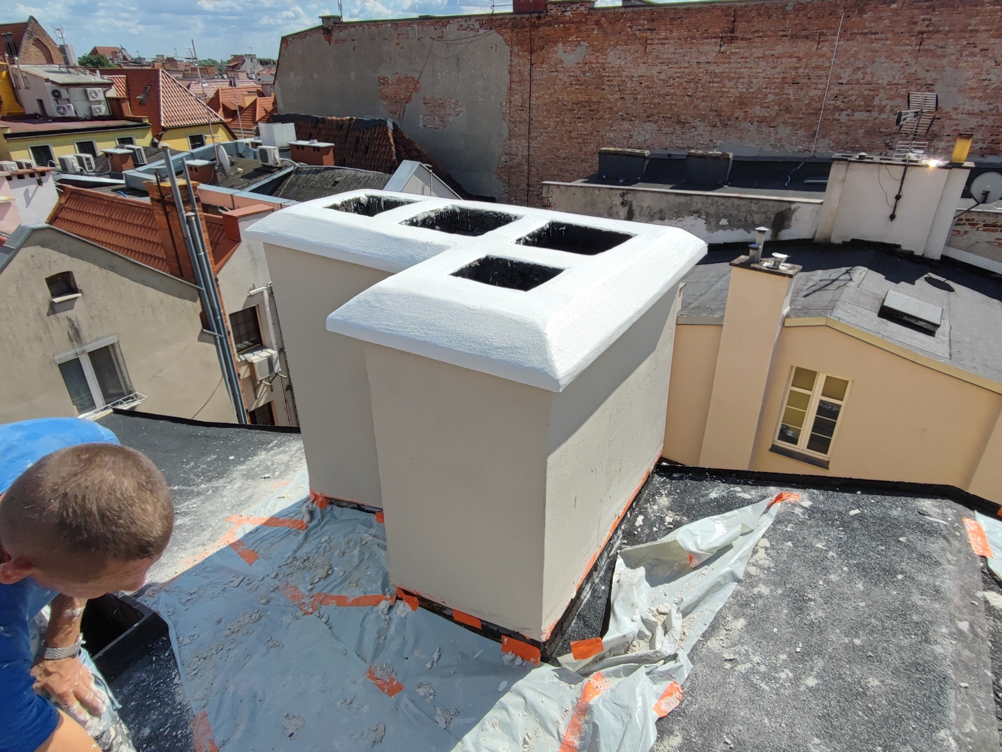 Prace na dachu: świeżo pomalowane kominy wentylacyjne z czterema otworami, zabezpieczone folią, widok z góry na tle miejskiej zabudowy w słoneczny dzień.