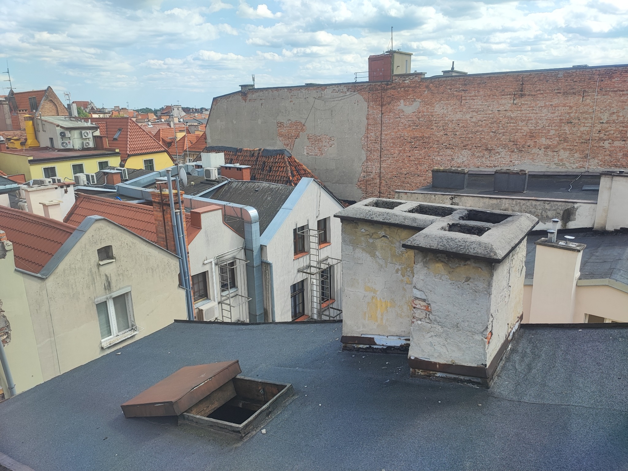 Widok z góry na dachy budynków w starej zabudowie miejskiej, z widocznymi kominami, oknem dachowym i fragmentami elewacji w tle.