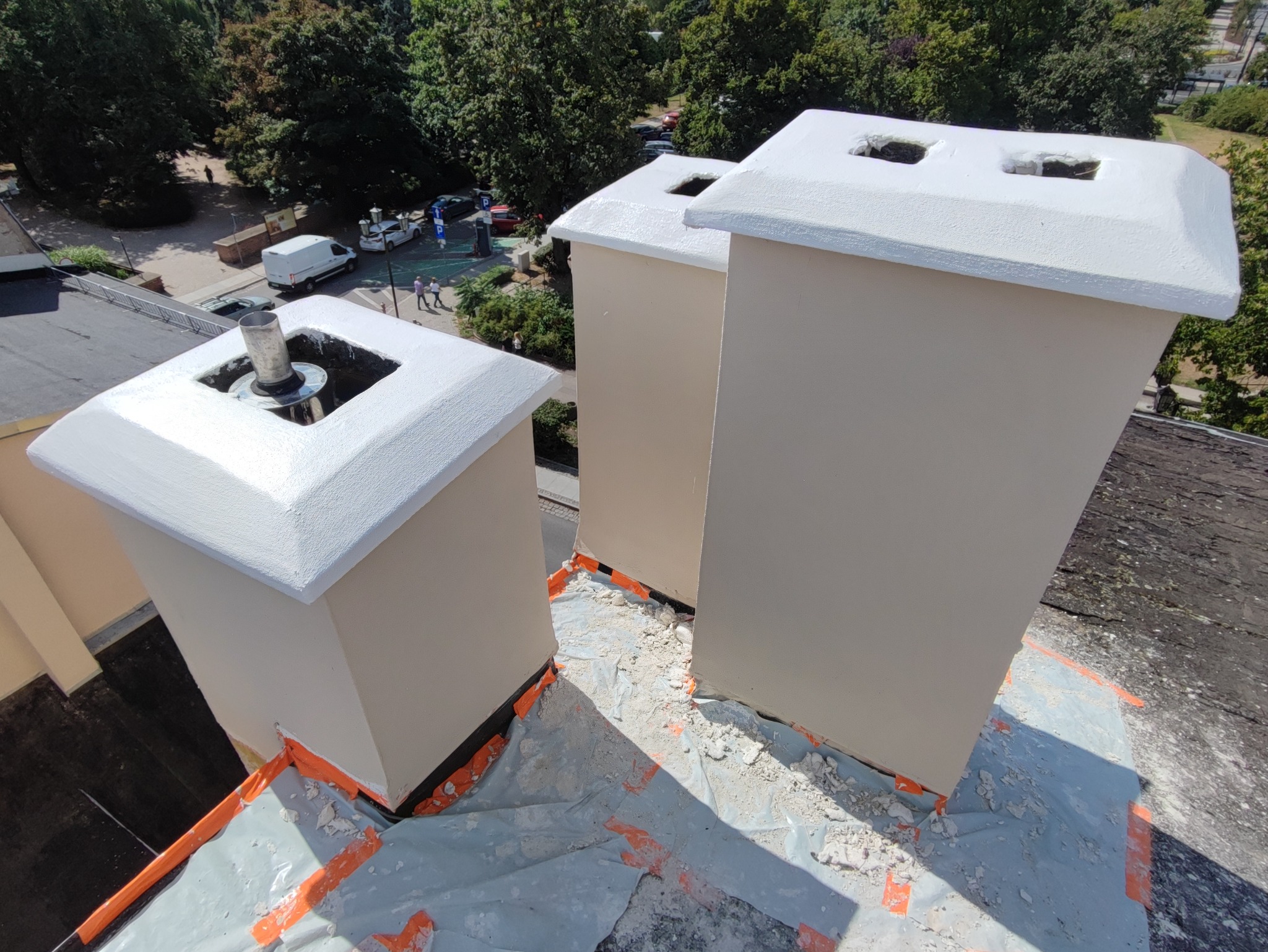 Trzy pomalowane kominy na dachu, zabezpieczone folią i taśmą malarską, widok z góry na tle drzew i ulicy.