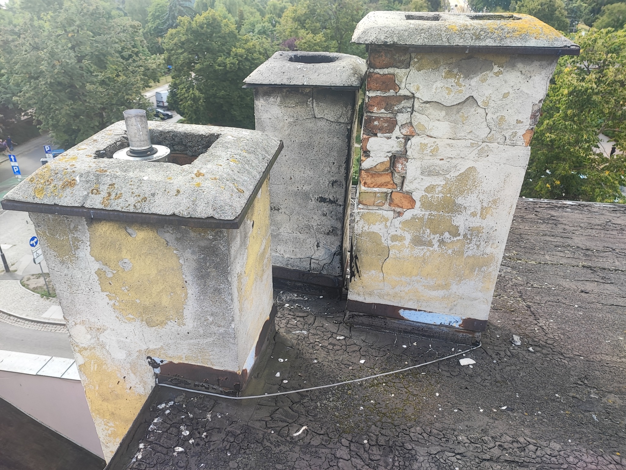 Trzy kominy na dachu, jeden z metalowym wkładem, z widocznymi uszkodzeniami tynku i cegieł, zarysowania farby, widoczne pęknięcia na dachu i mech, w tle drzewa i ulica.