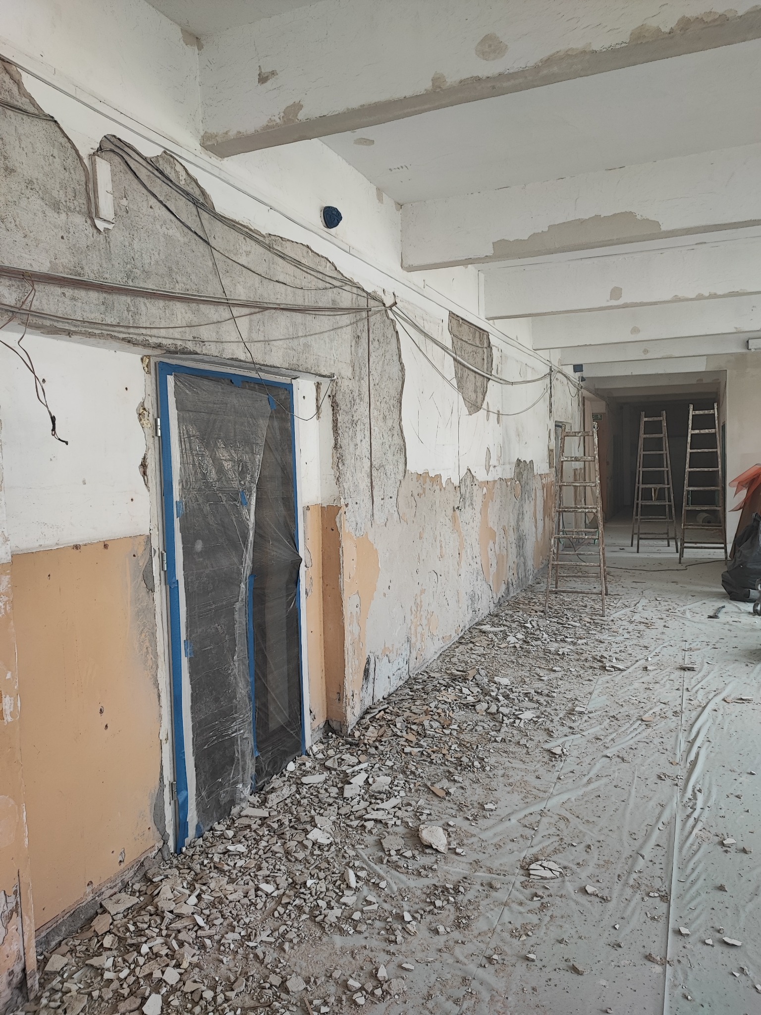 Wnętrze w trakcie remontu: fragment ściany z odsłoniętym murem, zabezpieczone folią drzwi z niebieską ramą, gruz na podłodze, drabiny w tle.