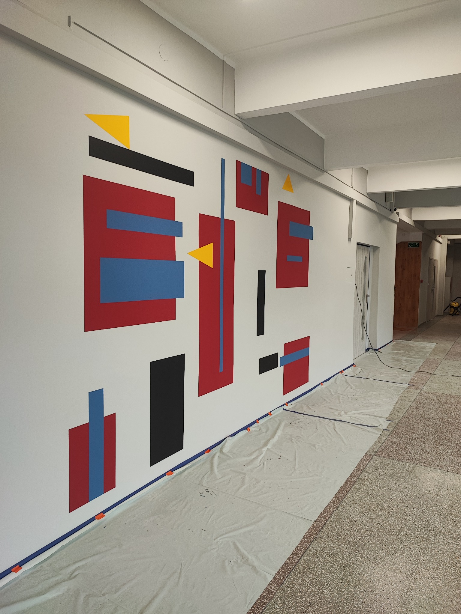 Wnętrze korytarza z geometryczną kompozycją malarską na ścianie, zabezpieczoną folią malarską i taśmą, widok z boku.