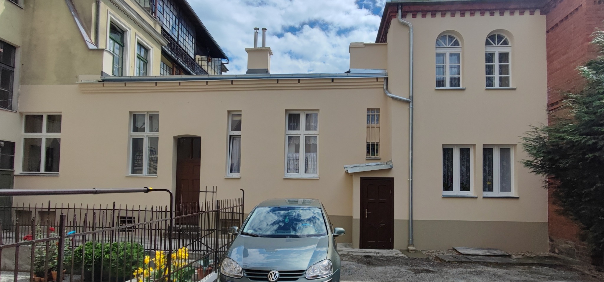 Odnowiona elewacja budynku z beżową fasadą, widoczne okna z białymi ramami, brązowe drzwi wejściowe, rynna, fragment dachu z kominem oraz zaparkowany samochód osobowy z odbiciem chmur w szybie.
