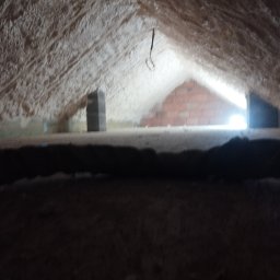 Usługi budowlane Krzysztof Bober - Poddasze po ociepleniu pianką PUR, widoczne ściany i dach pokryte grubą warstwą pianki, fragment muru z cegły w tle, ciemny element konstrukcyjny na pierwszym planie.