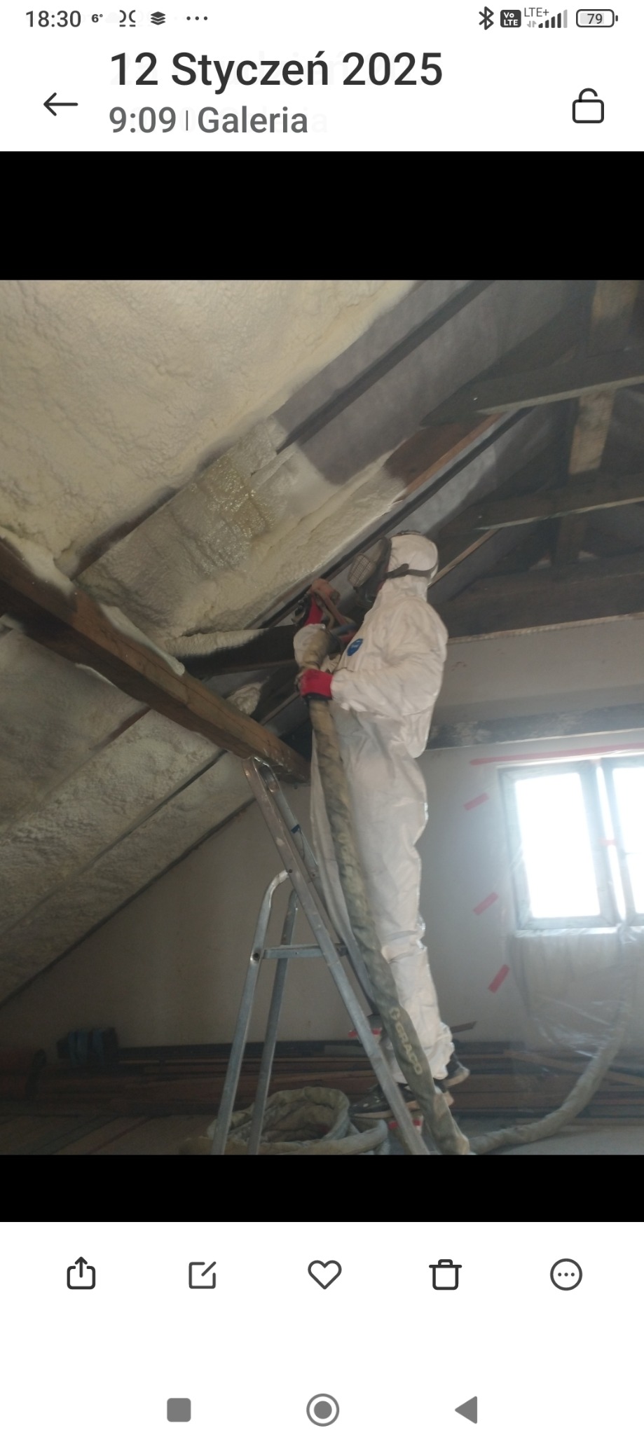 Pracownik w kombinezonie ochronnym i masce na drabinie, aplikuje piankę izolacyjną na poddaszu, widoczne drewniane elementy konstrukcyjne dachu i okno z folią ochronną.