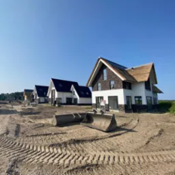 Nowo wybudowane domy jednorodzinne z oryginalnymi dachami, widoczne ślady prac ziemnych i łyżka koparki na pierwszym planie.