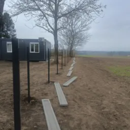 Etap budowy ogrodzenia: czarne metalowe słupki i betonowe podmurówki ułożone wzdłuż linii przyszłego ogrodzenia na tle pola i kontenera biurowego.