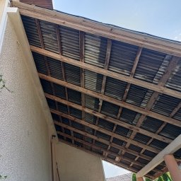 RAFBUD - Zewnętrzna elewacja domu z drewnianą konstrukcją zadaszenia, widok od dołu. Stara blacha falista na dachu, jasna elewacja, fragment zieleni.