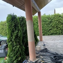 RAFBUD - Przygotowanie kolumny na zewnątrz budynku do malowania, z nałożoną warstwą szpachli, narzędziami i zabezpieczeniem podłoża folią. Widoczne tuje i kostka brukowa.