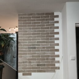 RAFBUD - Fragment ściany z dekoracyjną okładziną imitującą cegłę w odcieniach szarości, z widoczną fugą. W tle fragment rośliny doniczkowej i oświetlenia.