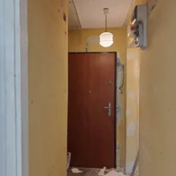 Korytarz w trakcie remontu: ściany z widocznymi śladami po szpachlowaniu, brązowe drzwi wejściowe, sufit z fakturą, wisząca lampa. Widoczne narzędzia i resztki materiał&oacute;w.