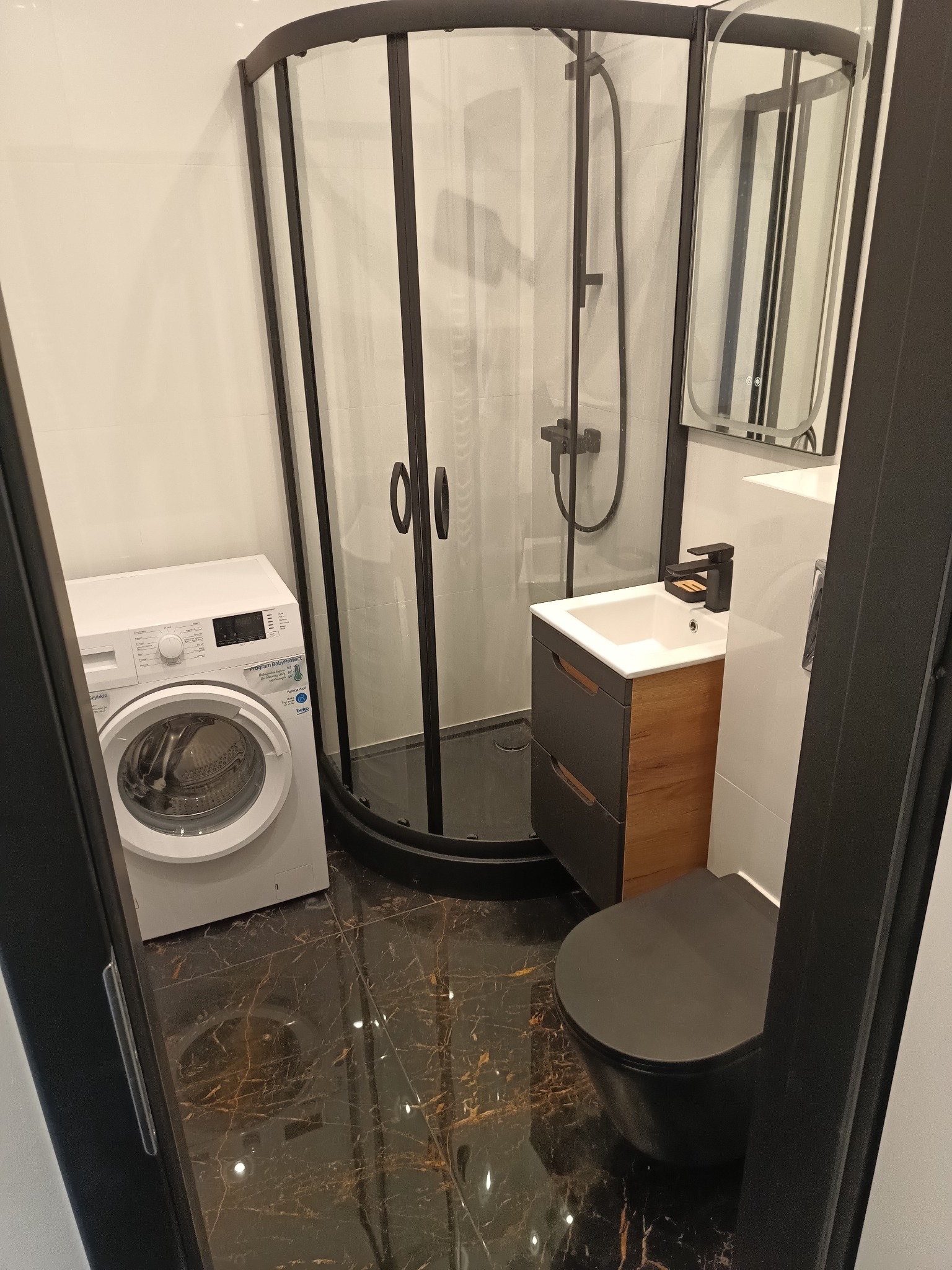 Nowoczesna łazienka z czarną armaturą i kabiną prysznicową, białą pralką oraz marmurową podłogą. Umywalka z szafką w kolorze drewna i czarną toaletą.