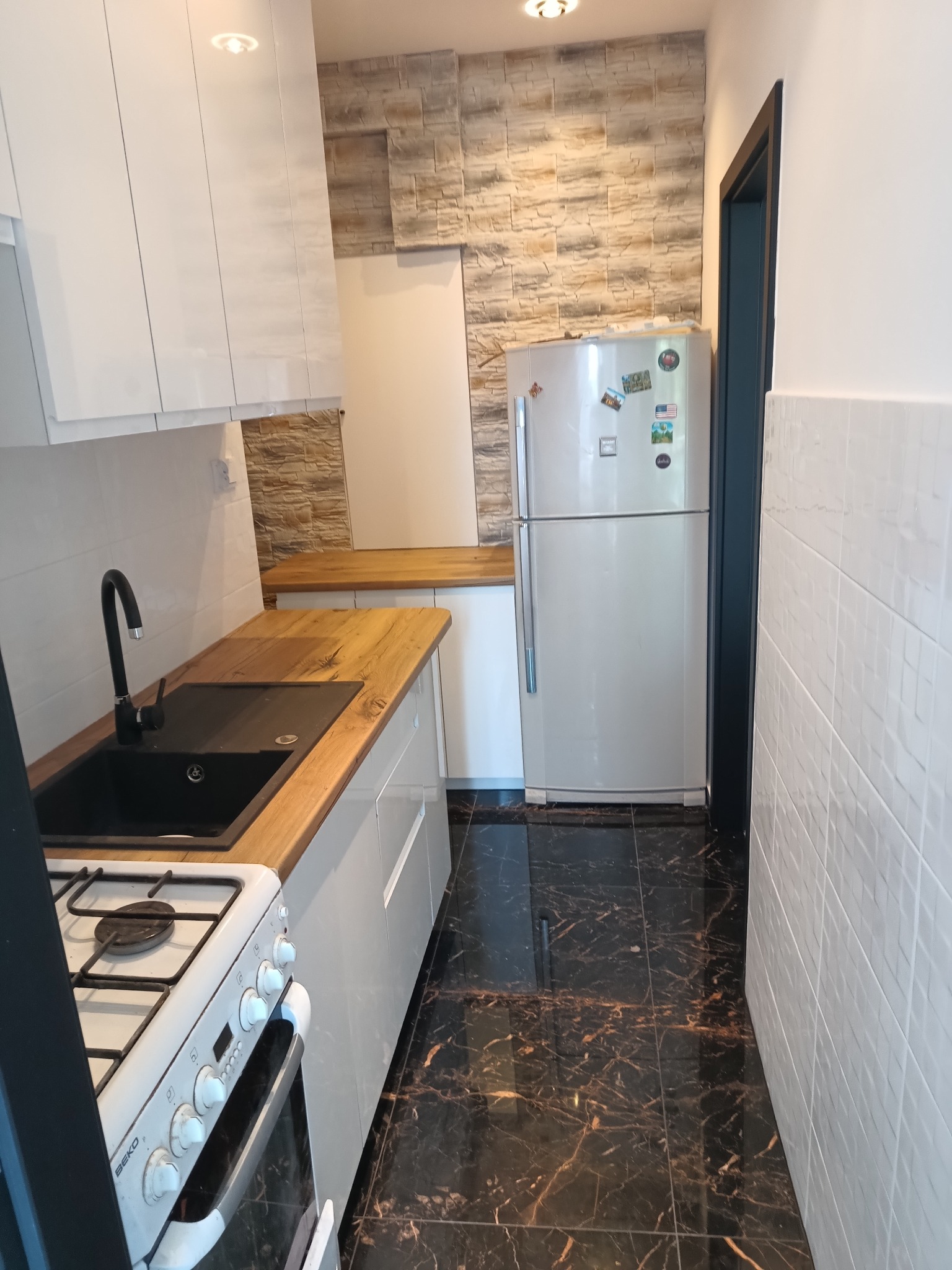 Nowoczesna kuchnia po remoncie: białe szafki z drewnianym blatem, czarny zlew, marmurowe płytki podłogowe i imitacja kamienia na ścianie. Widok na lodówkę.