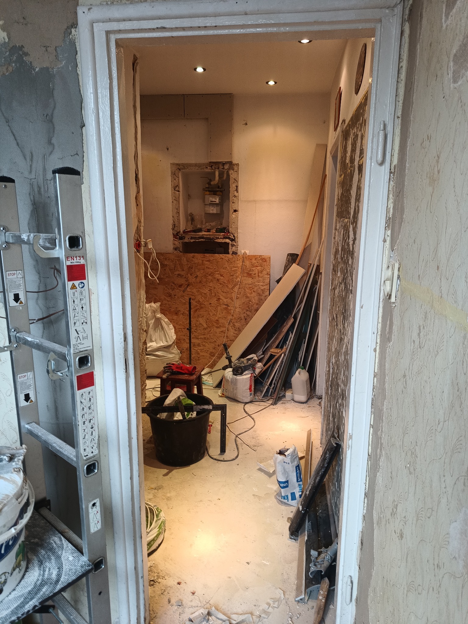 Remont wnętrza w trakcie: widoczna drabina, wiadro z zaprawą, płyta OSB, materiały budowlane i odsłonięte ściany. Ujęcie z perspektywy wejścia do pomieszczenia.