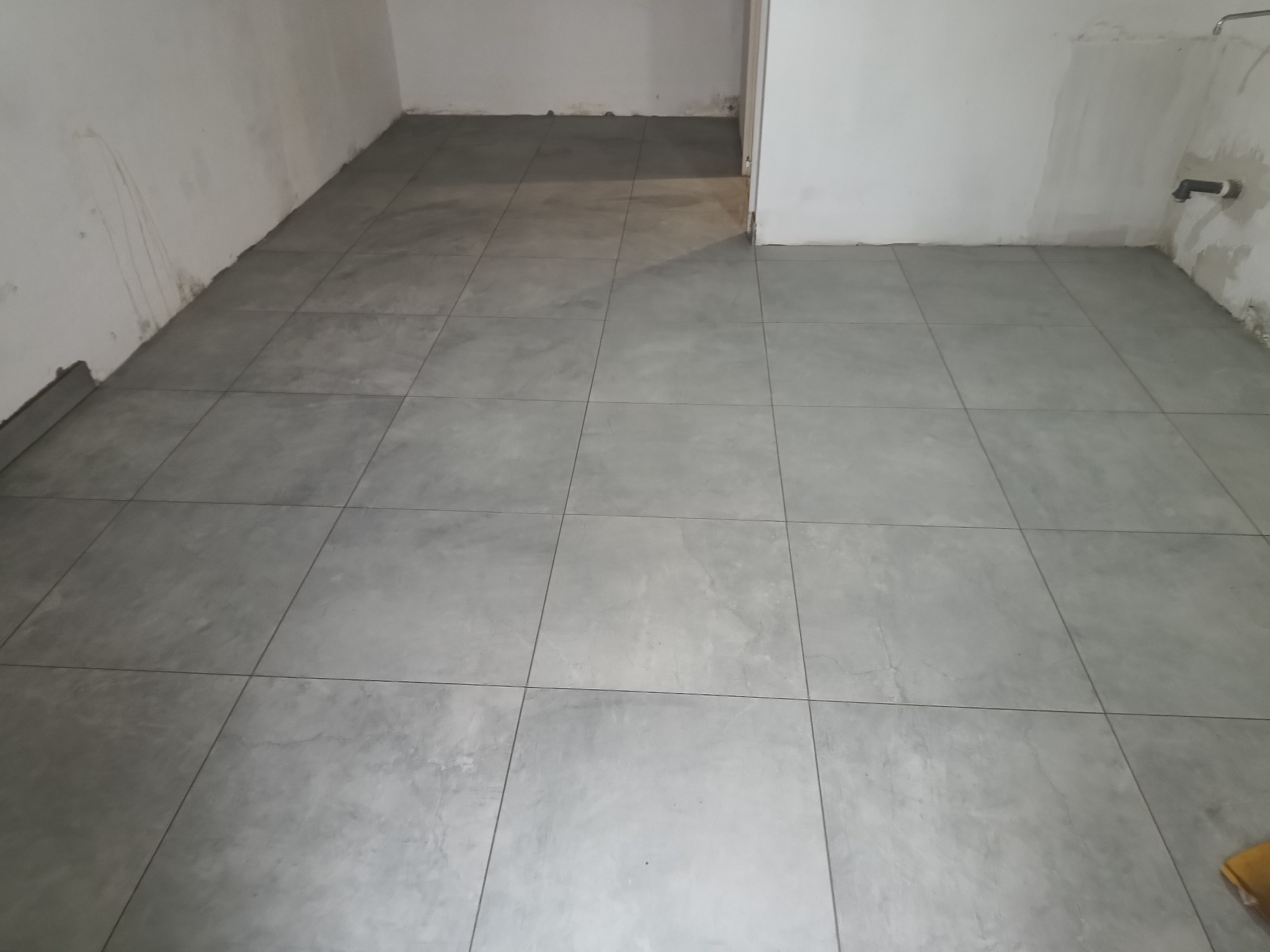 Ułożone szare płytki podłogowe w surowym wnętrzu. Widoczne niedoskonałości ścian i brak listew przypodłogowych. Minimalistyczny, industrialny styl.