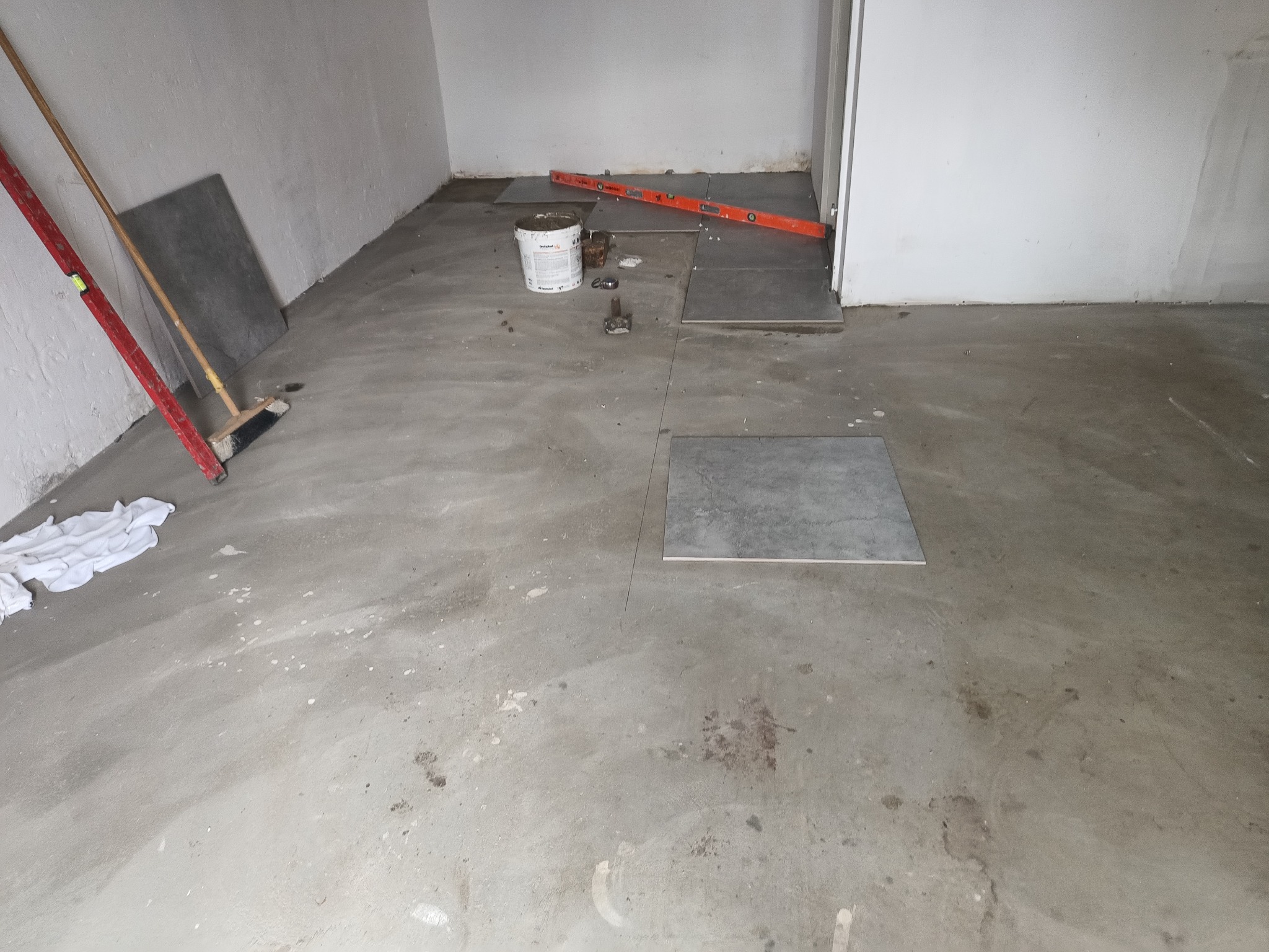 Wnętrze w trakcie układania szarych płytek podłogowych. Widoczny poziomica, wiadro z klejem i narzędzia glazurnicze na surowym, betonowym podłożu.