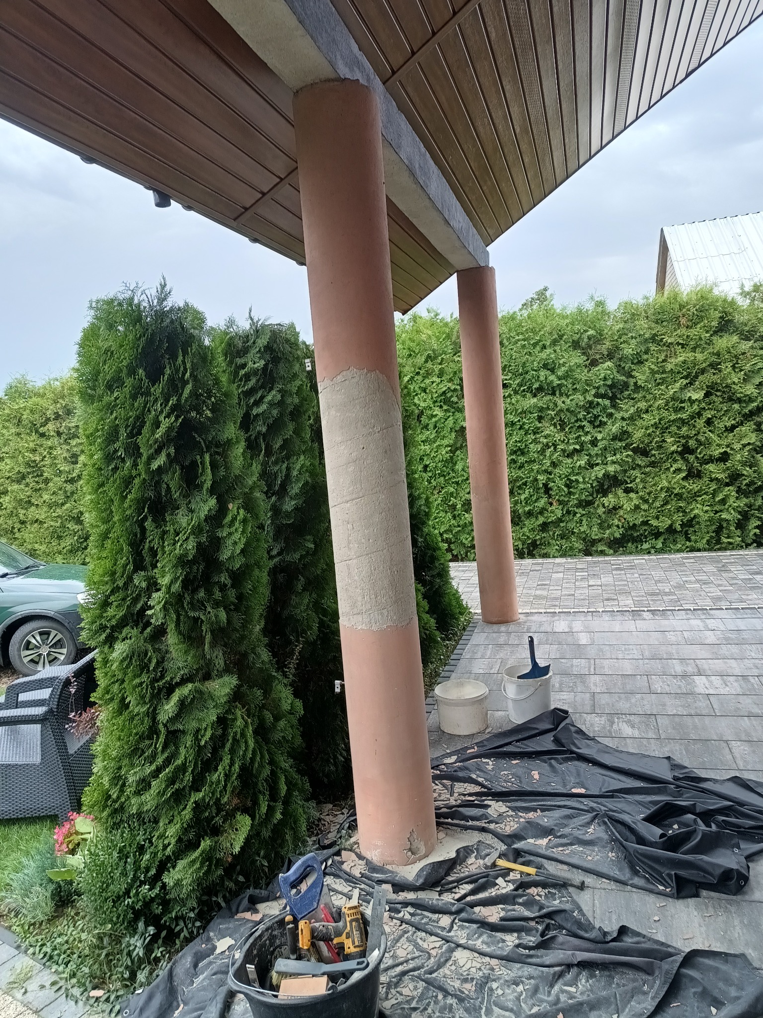 Przygotowanie kolumny na zewnątrz budynku do malowania, z nałożoną warstwą szpachli, narzędziami i zabezpieczeniem podłoża folią. Widoczne tuje i kostka brukowa.