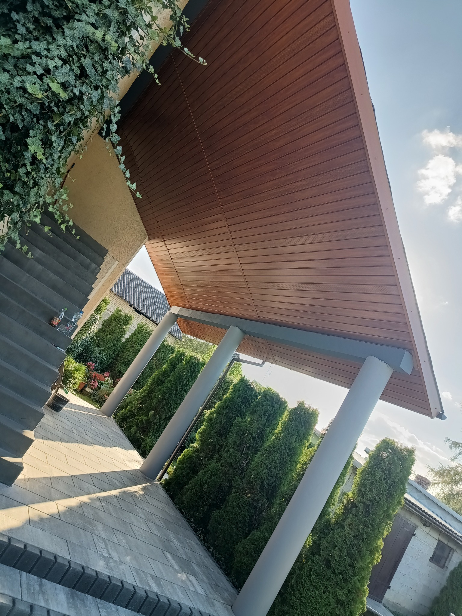 Zadaszenie tarasu z drewnianą boazerią i kolumnami, widok z dołu. Schody prowadzące do wejścia, dekoracyjne tuje wzdłuż ścieżki, błękitne niebo w tle.