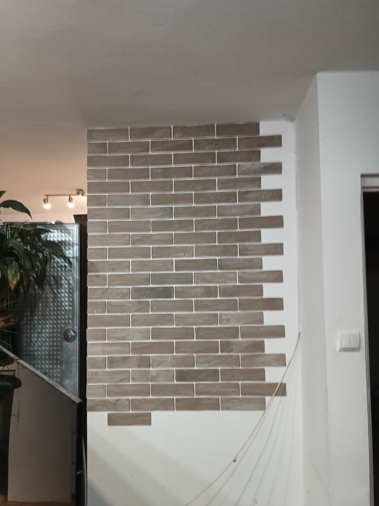 Fragment ściany z dekoracyjną okładziną imitującą cegłę w odcieniach szarości, z widoczną fugą. W tle fragment rośliny doniczkowej i oświetlenia.