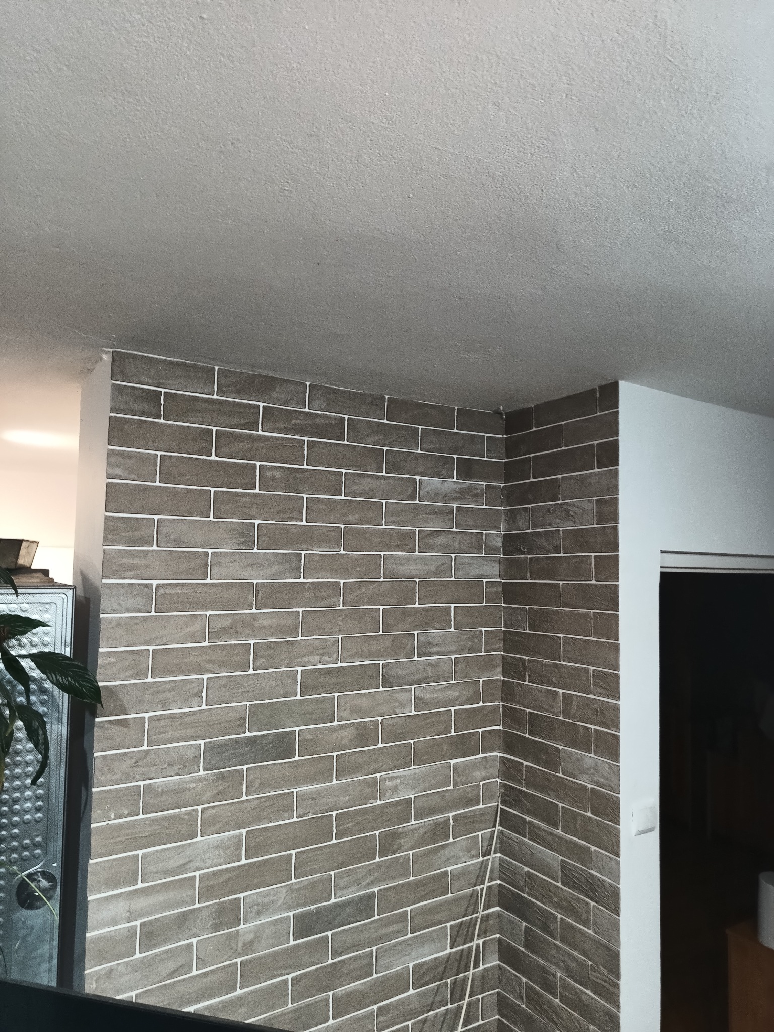 Ściana wykończona dekoracyjną cegłą w odcieniach szarości z białymi fugami, widoczna w rogu pomieszczenia, z fragmentem sufitu i drzwi po prawej stronie.