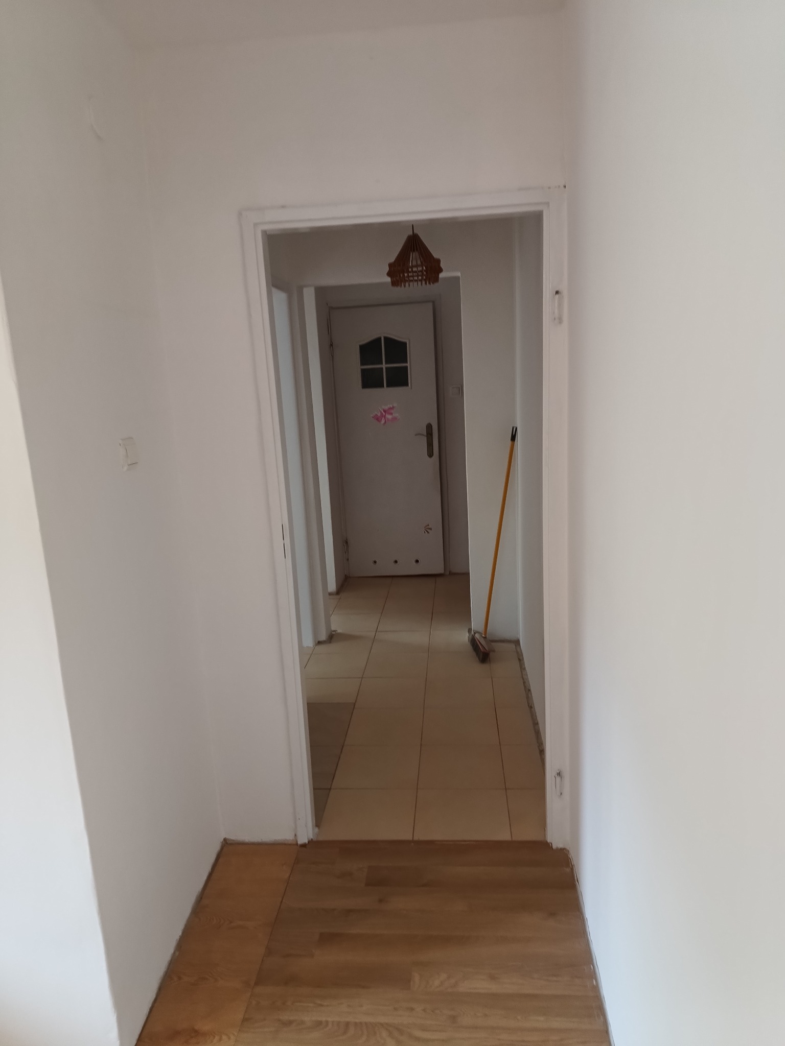 Białe ściany korytarza z nowo ułożonymi panelami podłogowymi w odcieniach brązu. W głębi widoczne drzwi wejściowe z dekoracją i lampa.