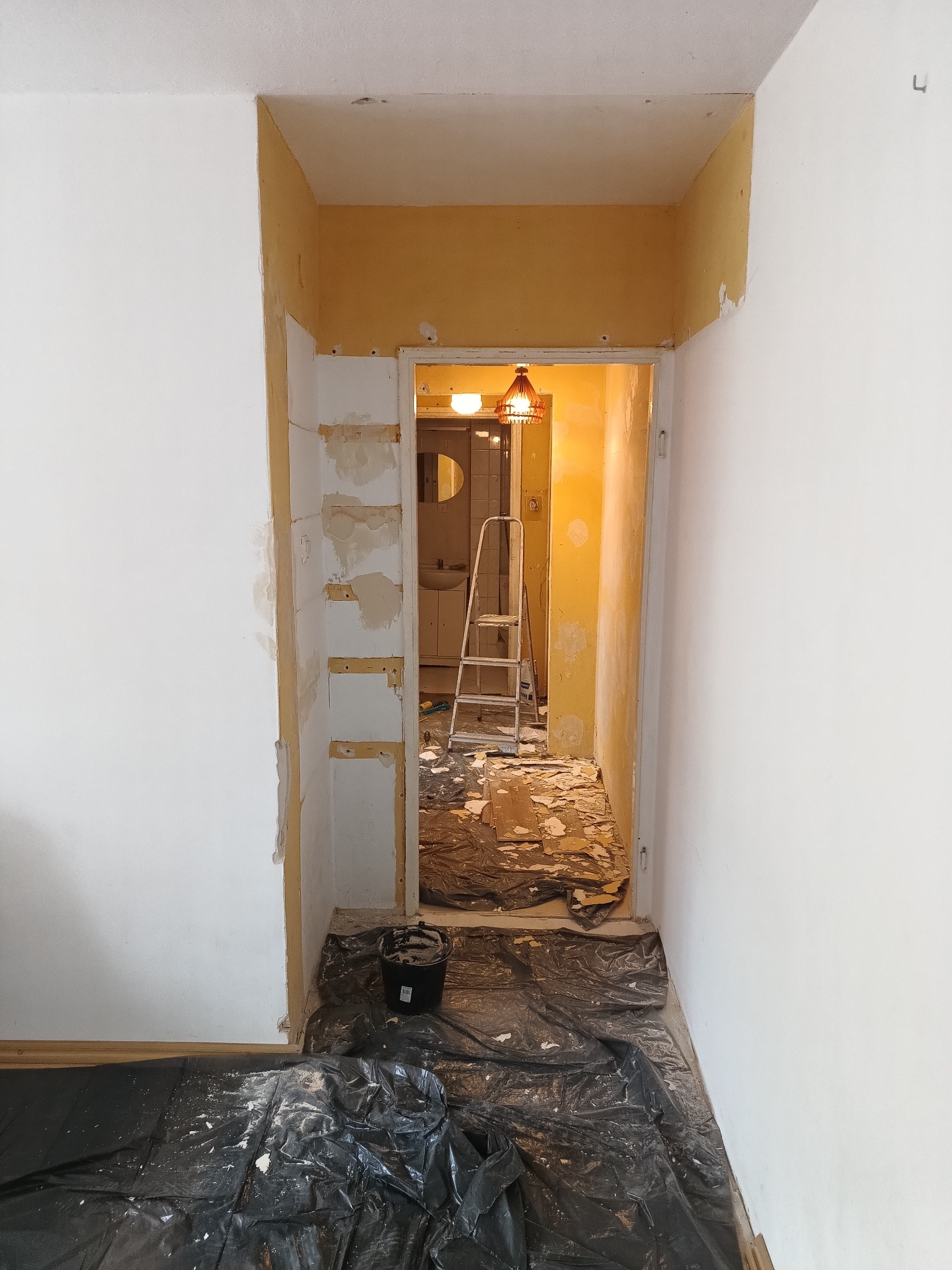 Remont mieszkania: przygotowanie ścian do malowania. Widoczne ślady po szpachlowaniu, folia ochronna na podłodze i drabina w przejściu do kolejnego pomieszczenia.