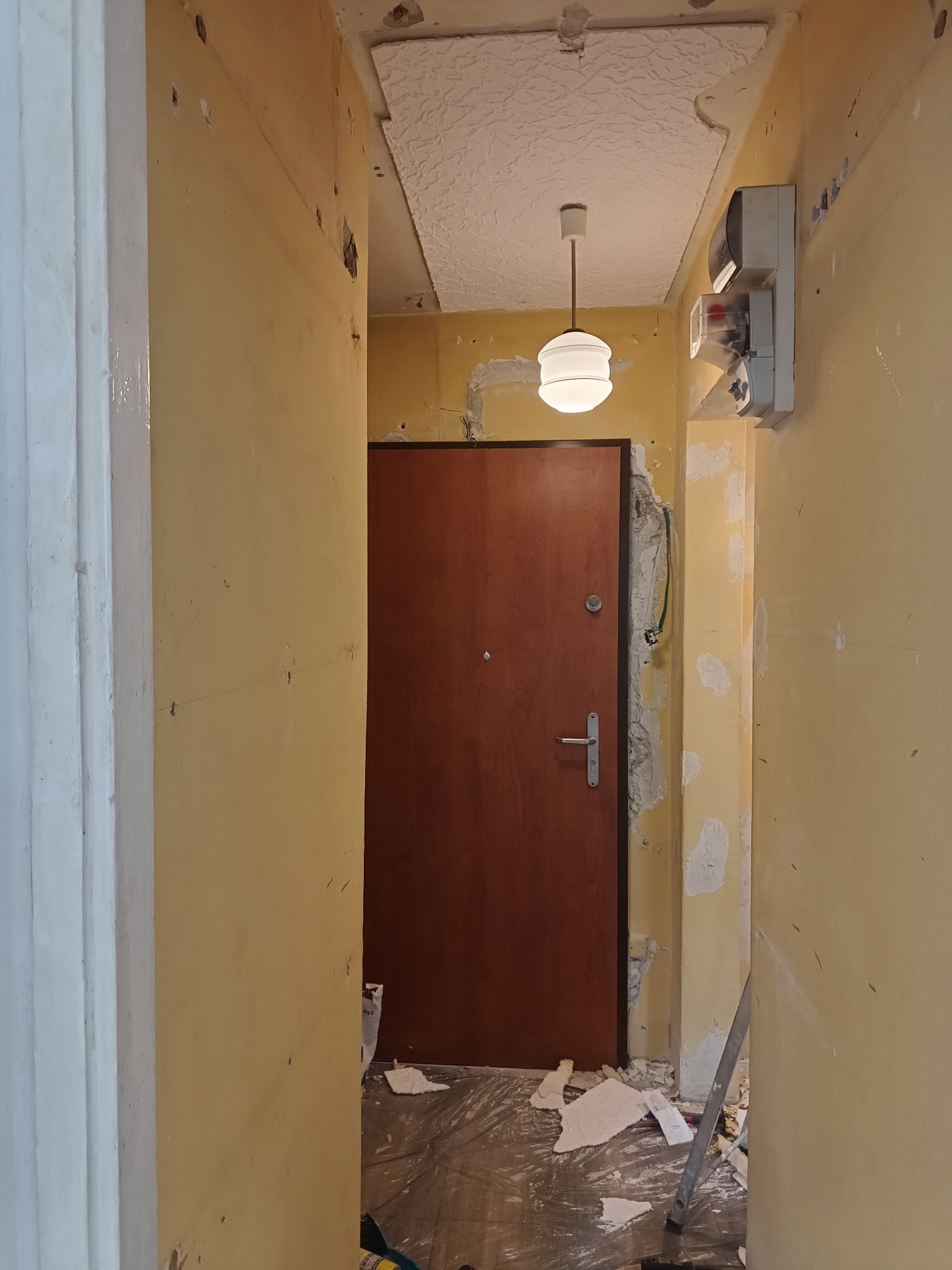 Korytarz w trakcie remontu: ściany z widocznymi śladami po szpachlowaniu, brązowe drzwi wejściowe, sufit z fakturą, wisząca lampa. Widoczne narzędzia i resztki materiałów.