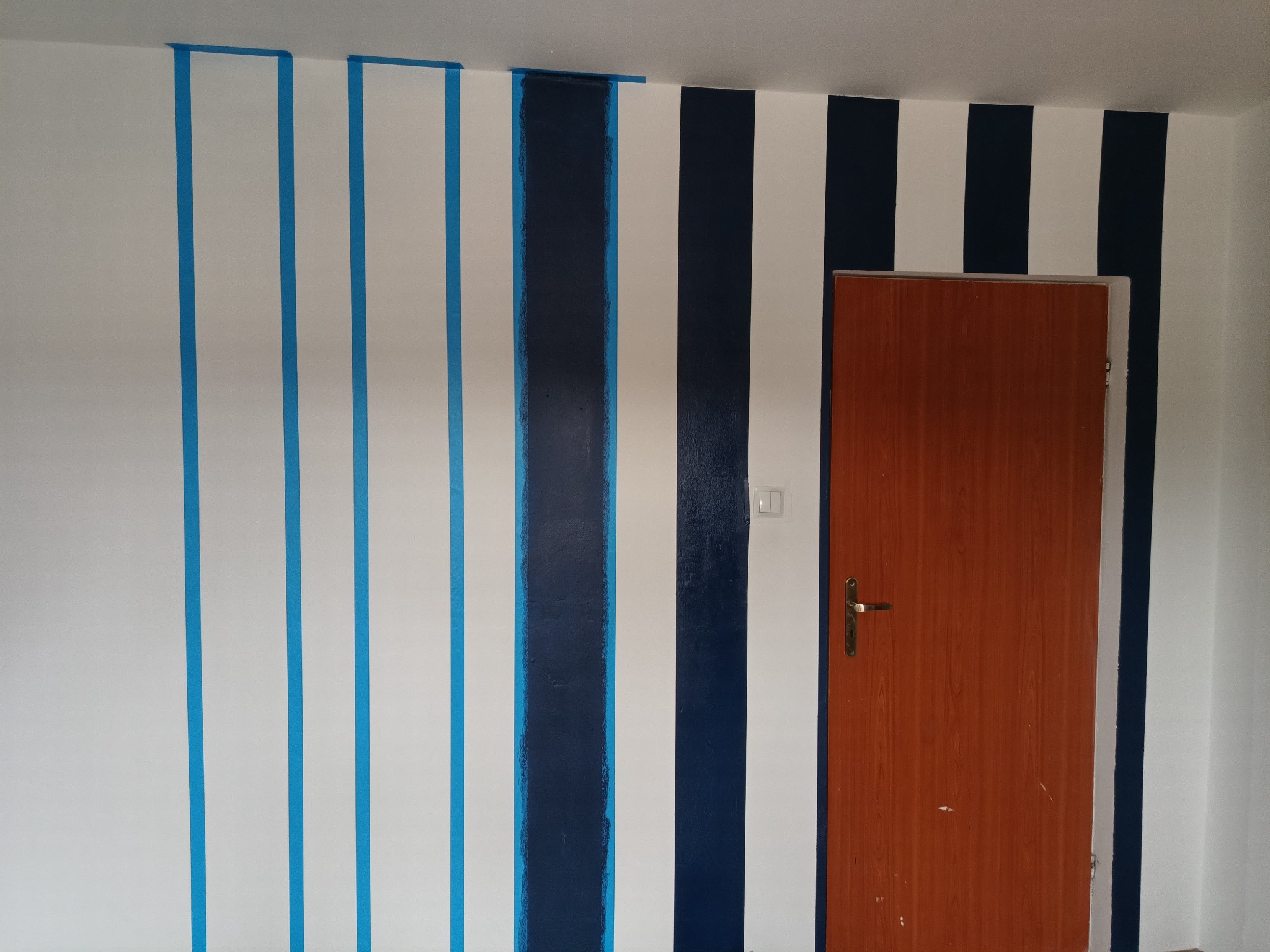 Ściana w pionowe pasy malowana na granatowo i biało, z taśmą malarską, w trakcie realizacji w Kozienicach. Widoczna część brązowych drzwi.
