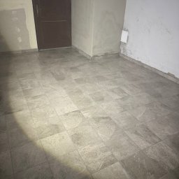 ZAKŁAD OGÓLNOBUDOWLANY MATEUSZ ZYGMOND - Wykończone podłogi z szarych płytek imitujących marmur w pomieszczeniu w trakcie remontu. Widoczne ślady po gładzi na ścianach i brązowe drzwi.