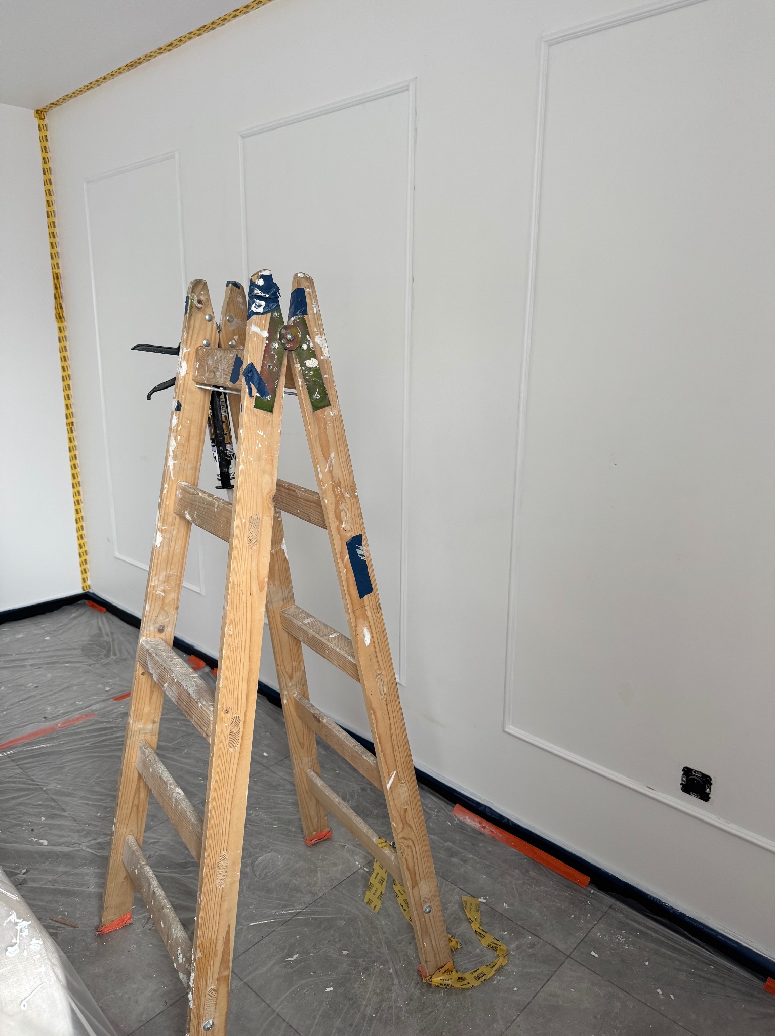 Białe ściany z dekoracyjnymi listwami, zabezpieczone taśmą malarską. Drewniana drabina malarska stoi na folii ochronnej. Przygotowanie do malowania.