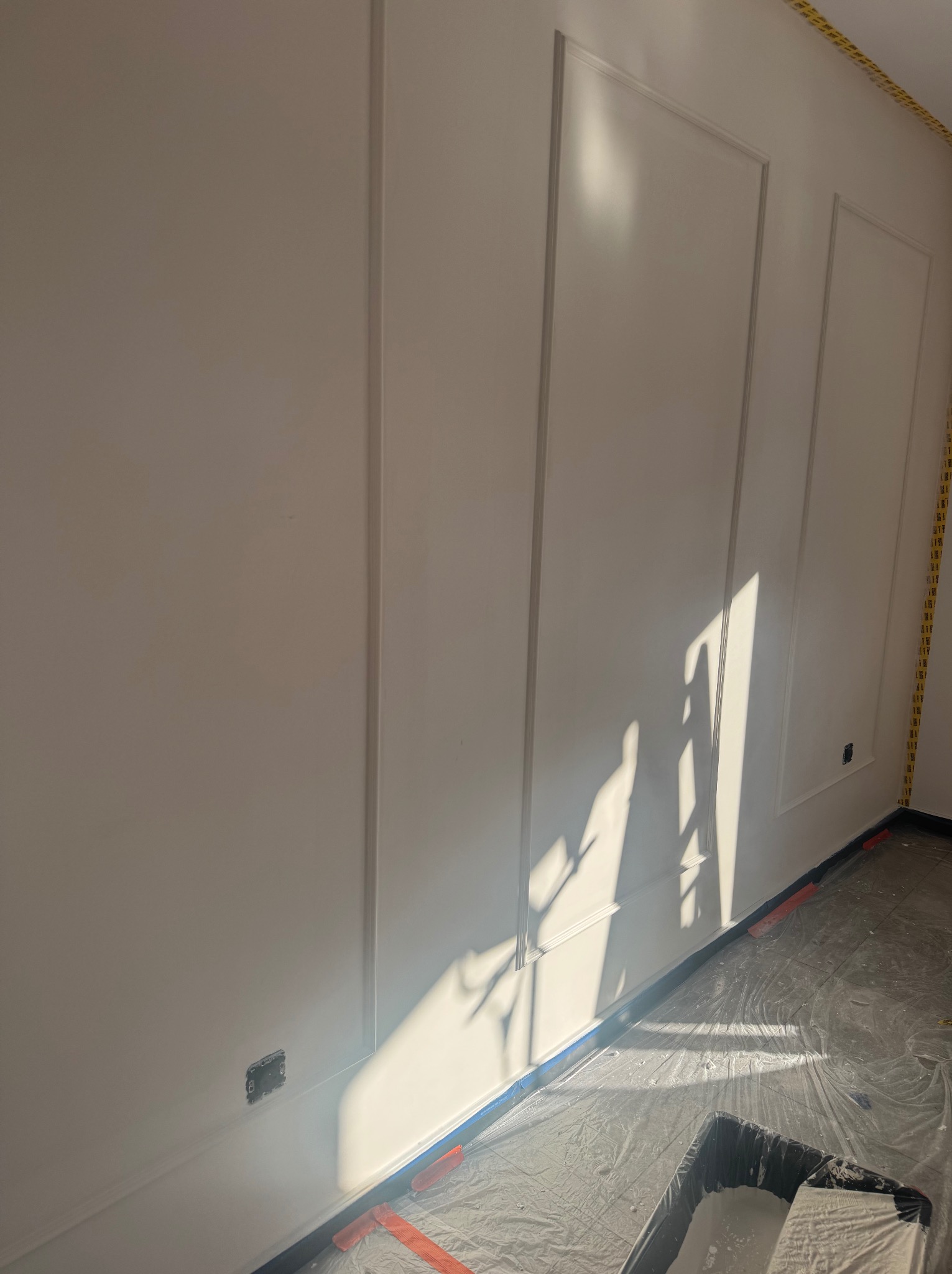 Biała ściana z dekoracyjnymi panelami w trakcie malowania, zabezpieczona folią i taśmą malarską, z widocznym wałkiem i kuwetą z farbą. Padające światło słoneczne.