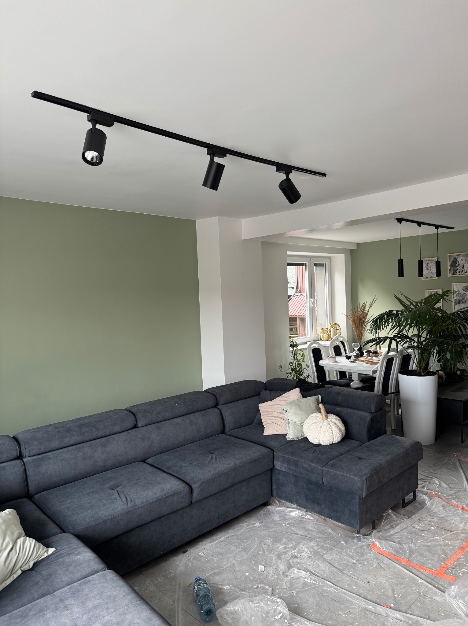 Salon po odświeżeniu: oliwkowa ściana, grafitowa sofa narożna z dekoracyjnymi poduszkami, czarne reflektory szynowe. Podłoga zabezpieczona folią malarską.
