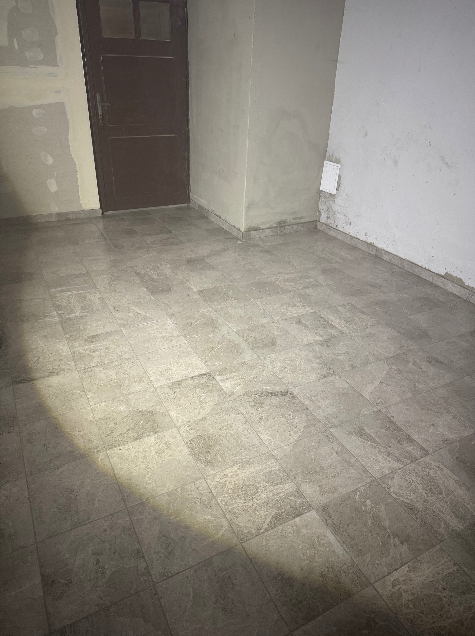 Wykończone podłogi z szarych płytek imitujących marmur w pomieszczeniu w trakcie remontu. Widoczne ślady po gładzi na ścianach i brązowe drzwi.