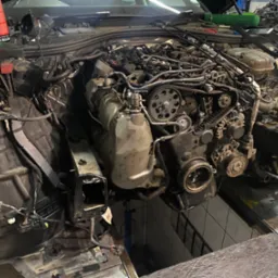 Rozłożony silnik samochodowy w warsztacie, widoczne elementy układu rozrządu i osprzętu, brak przedniego pasa. Auto na podnośniku.