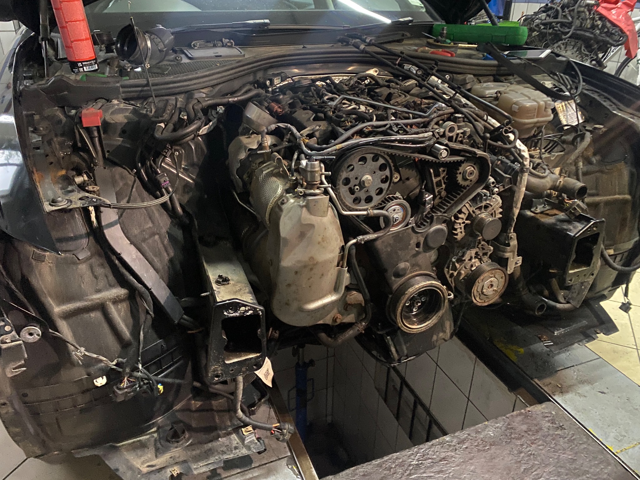 Rozłożony silnik samochodowy w warsztacie, widoczne elementy układu rozrządu i osprzętu, brak przedniego pasa. Auto na podnośniku.