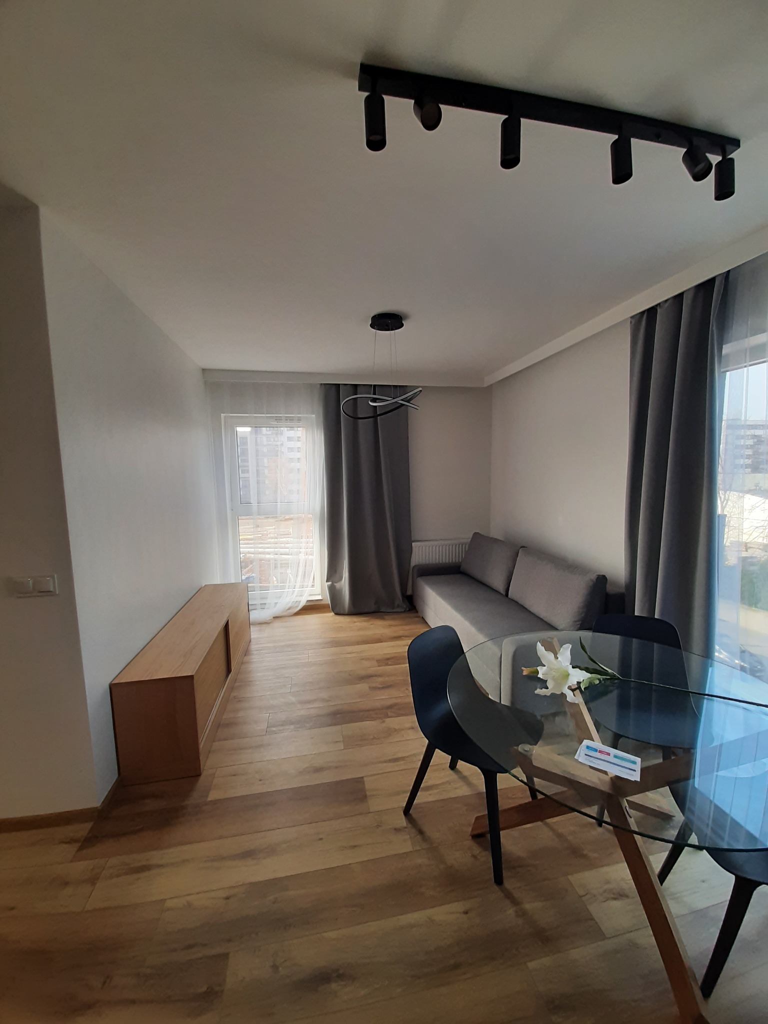 Jasne wnętrze pokoju dziennego z drewnianą podłogą, szarą sofą, szarymi zasłonami, szklanym stołem z czarnymi krzesłami i bukietem białych kwiatów na stole, oświetlenie sufitowe punktowe.