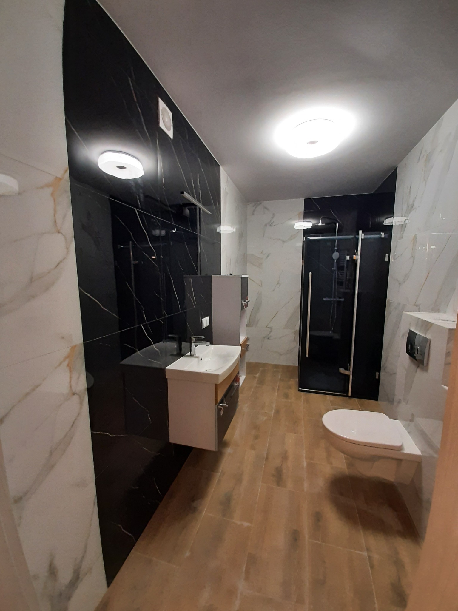 Nowoczesna łazienka z czarno-białymi marmurowymi płytkami, drewnopodobną podłogą i kabiną prysznicową typu walk-in z czarnymi profilami.