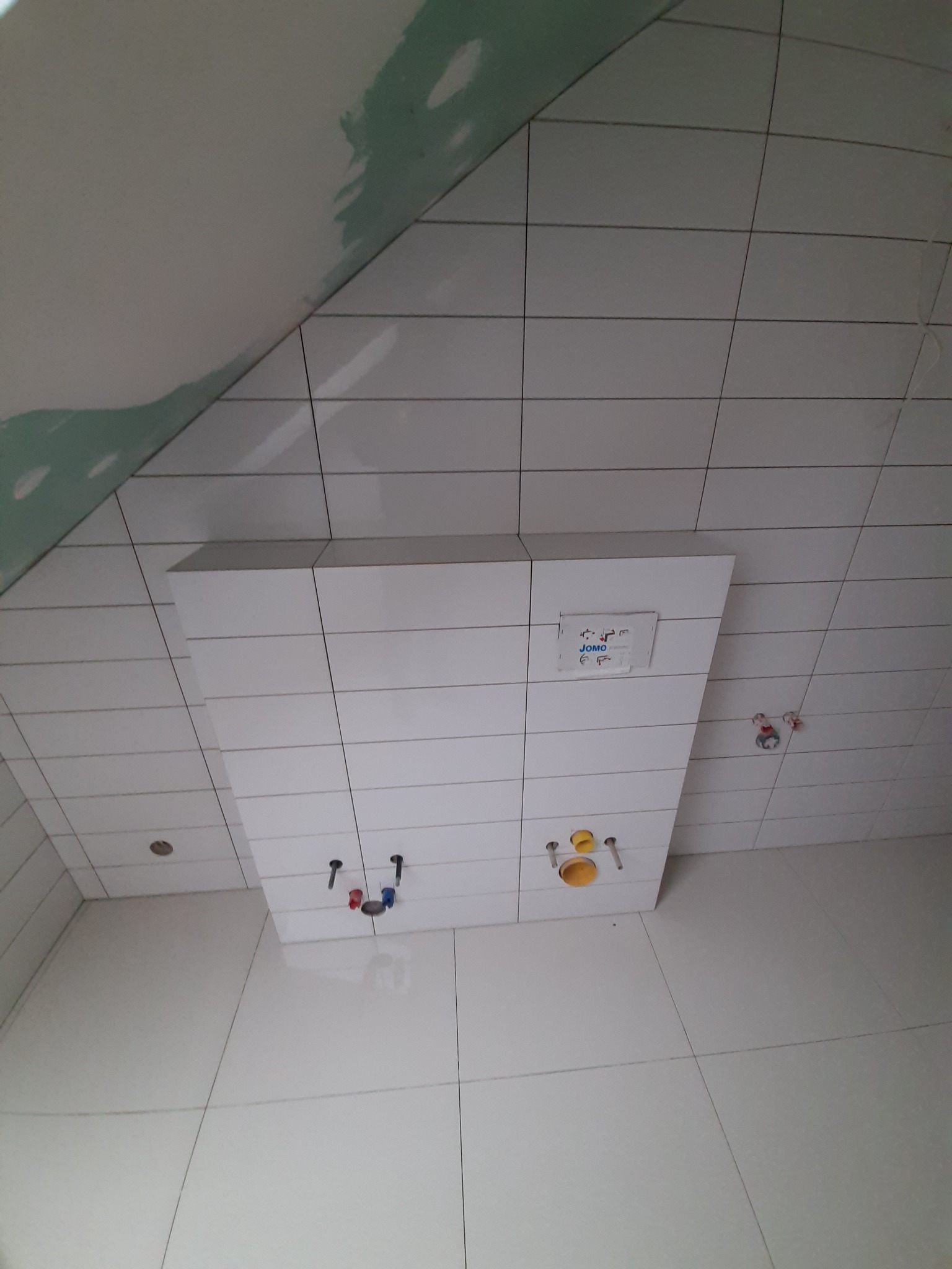 Białe płytki w łazience na poddaszu, widoczna zabudowa stelaża podtynkowego WC z wystającymi rurami instalacyjnymi, ściana częściowo przygotowana pod malowanie.