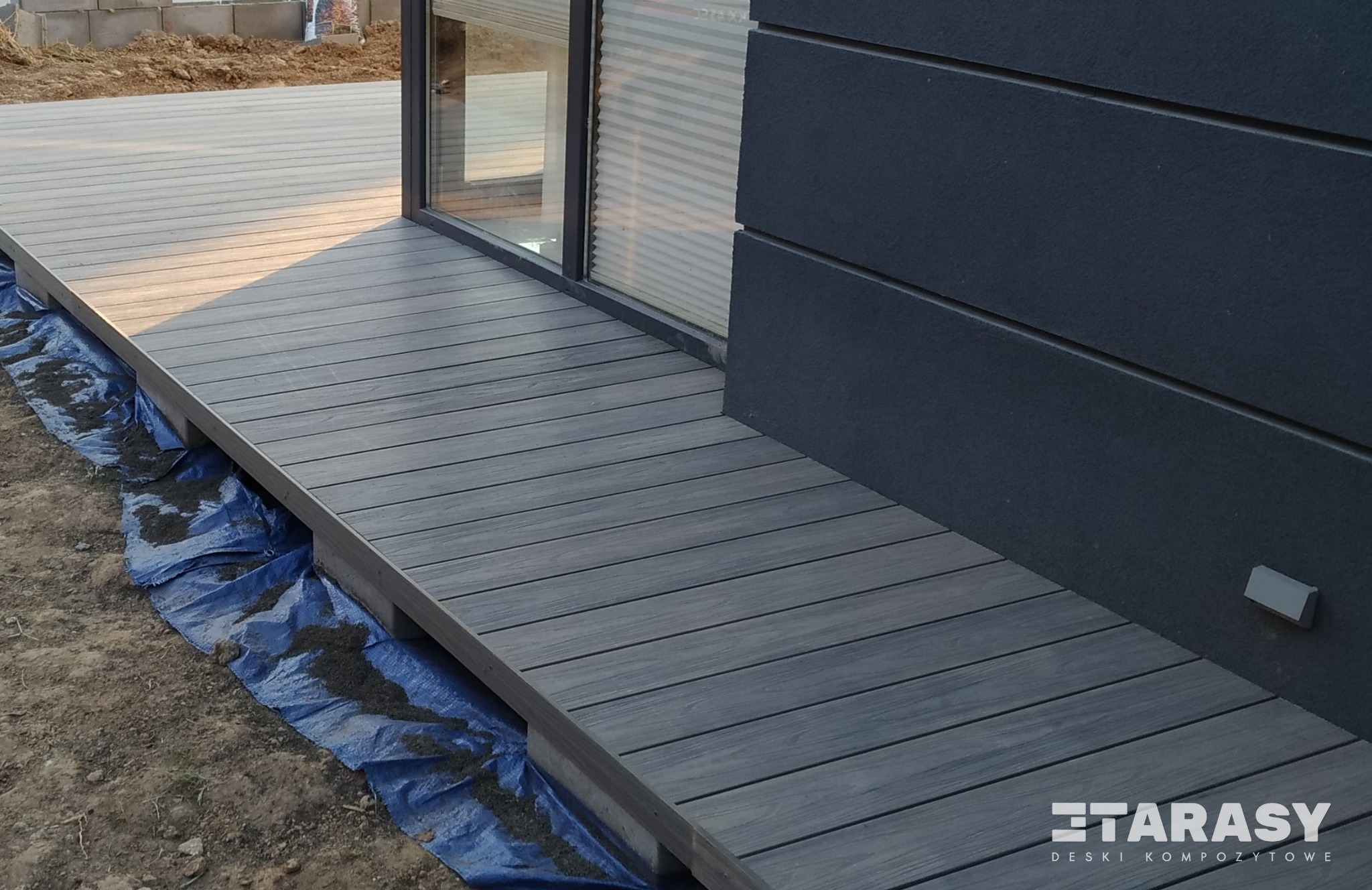 Szary taras z desek ułożonych przy ścianie budynku, widoczna niebieska folia pod konstrukcją, okno z żaluzjami w tle, ziemia wokół, w prawym dolnym rogu logo producenta.