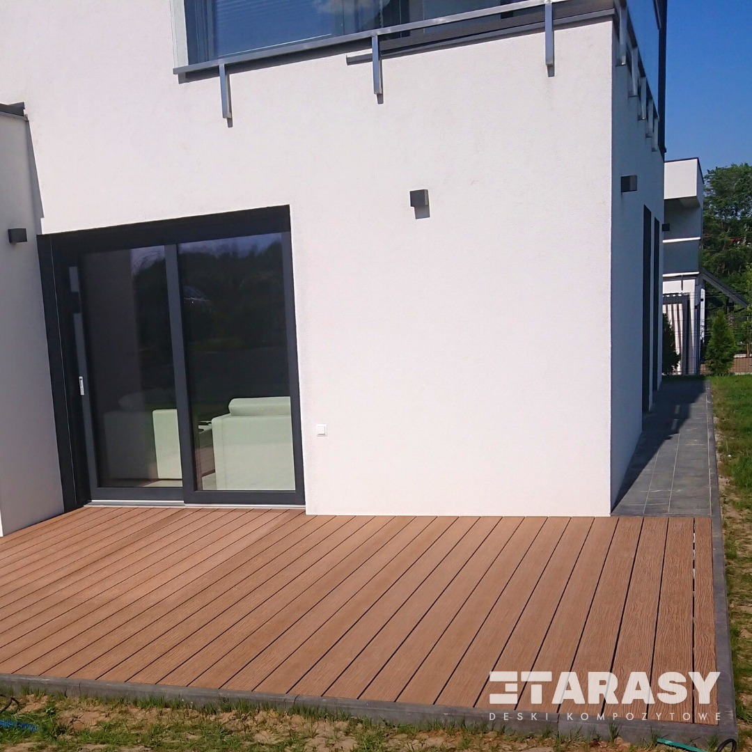 Nowoczesny taras z desek w kolorze jasnego drewna, przylegający do białego budynku z dużym przesuwnym oknem. Widoczna trawa i szary chodnik.