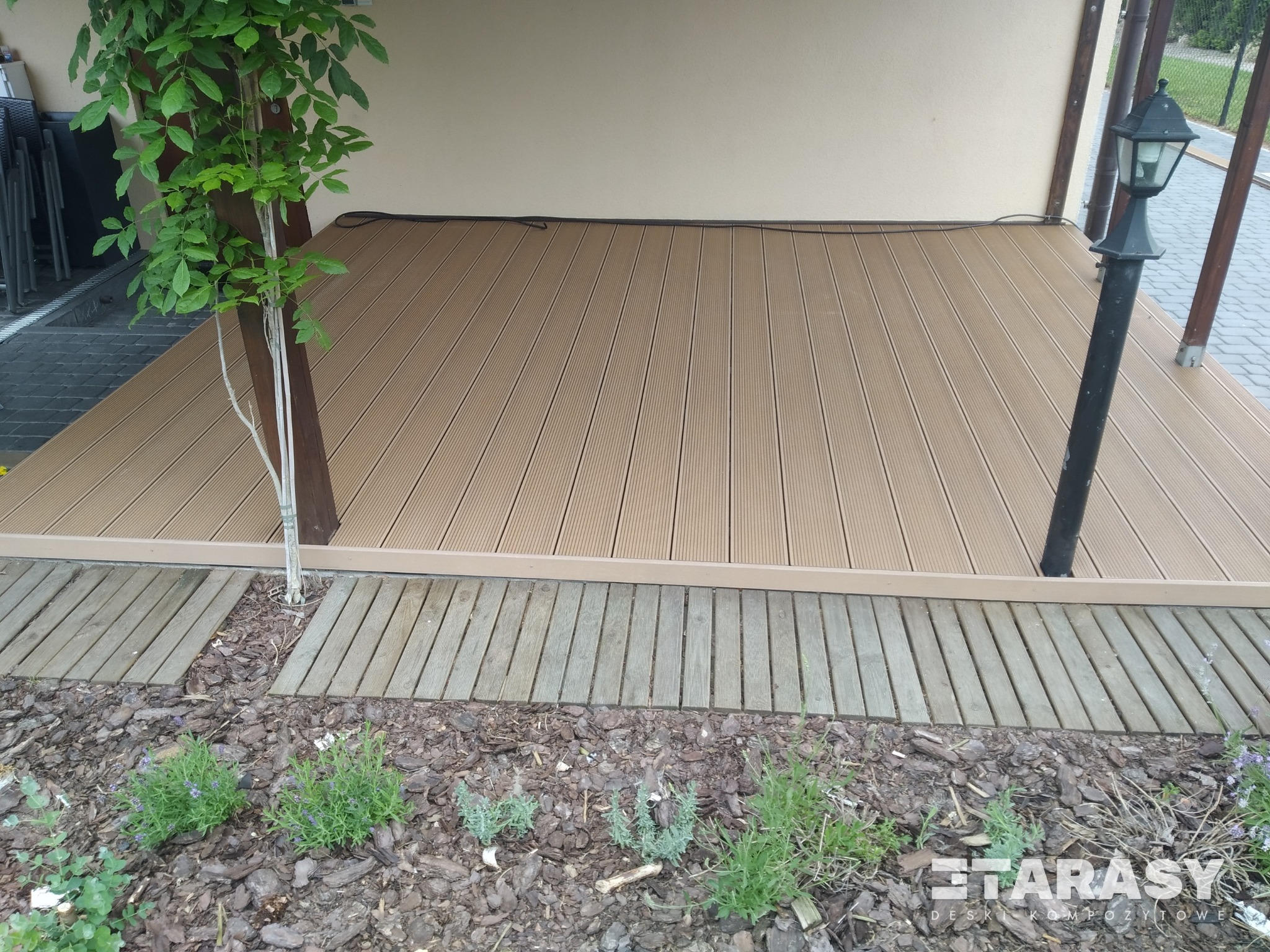 Beżowy taras kompozytowy z oświetleniem zewnętrznym i obrzeżem z desek, otoczony korą i niską roślinnością.