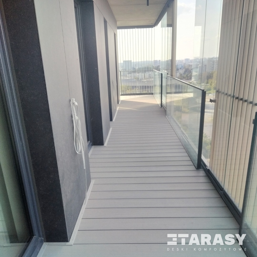 Szary taras na balkonie z deskami kompozytowymi i szklaną balustradą, widok na miasto w tle, logo producenta desek w prawym dolnym rogu.