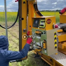 Operator w czerwonych rękawicach obsługuje żółtą maszynę Wamet do wbijania słupków ogrodzeniowych na tle kwitnącego pola rzepaku.